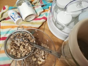 INGREDIENTES YOGUR VEGANO EN YOGURTERA Sobre una mesa yogurtera con yogures rodeado de semillas un bote de almidón y otro de probióticos sobre un trapo de colores