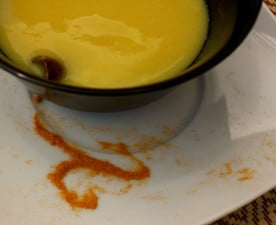 sobre un plato blanco hay una taza negra que contiene leche de quinoa con color de cúrcuma. un poco de especia de cúrcuma adorna la escena