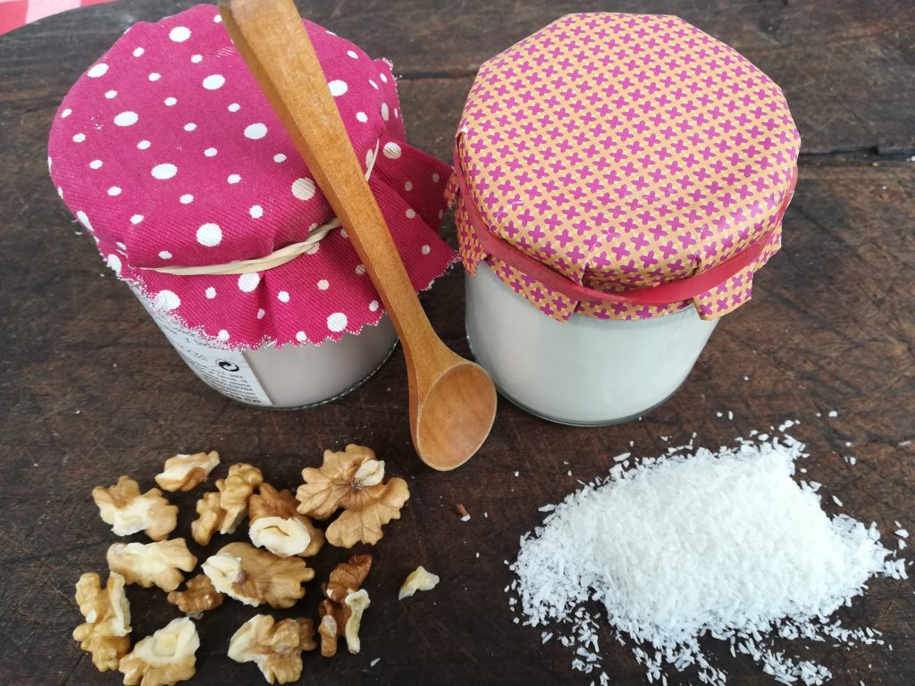 receta vegana de yogur de coco en yogurtera dos tarros de vidrio con yogur sobre la mesa. Coco rayado y nueces al rededor